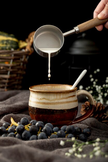Equipaggi il latte di versamento in una tazza di caffè marrone con l'uva su un panno grigio