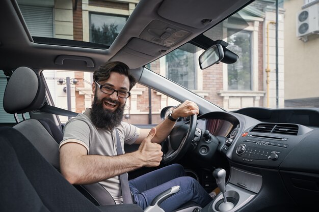 Equipaggi l'autista sorridente felice che mostra i pollici su che conducono l'automobile sportiva