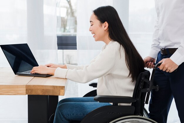 Equipaggi la condizione dietro la donna disattivata sorridente che si siede sulla sedia a rotelle facendo uso del computer portatile