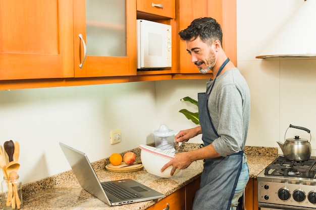 Equipaggi la preparazione del cibo guardando la ricetta sul computer portatile nella cucina