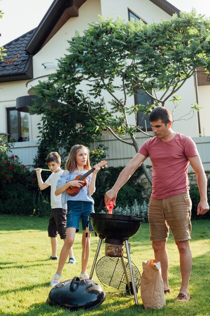 Equipaggi la preparazione dell&#39;alimento sulla griglia del barbecue mentre sua figlia che gioca le ukulele