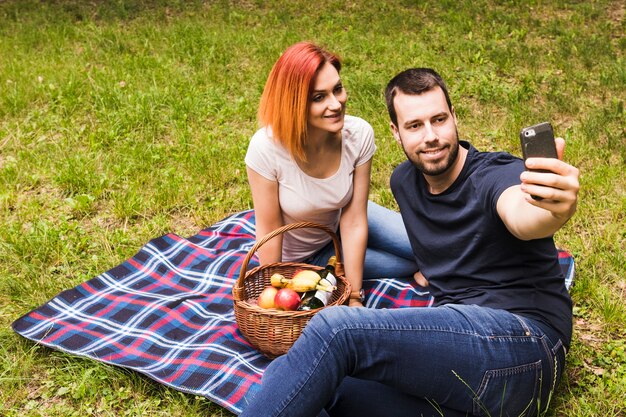 Equipaggi la presa del selfie con la sua ragazza sorridente sul telefono cellulare al picnic