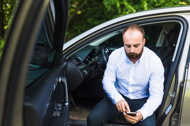 Equipaggi la seduta in un&#39;automobile con la porta aperta che esamina lo schermo del telefono cellulare