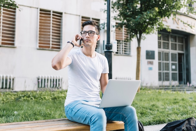 Equipaggi la seduta sul banco con il computer portatile che parla sul cellulare