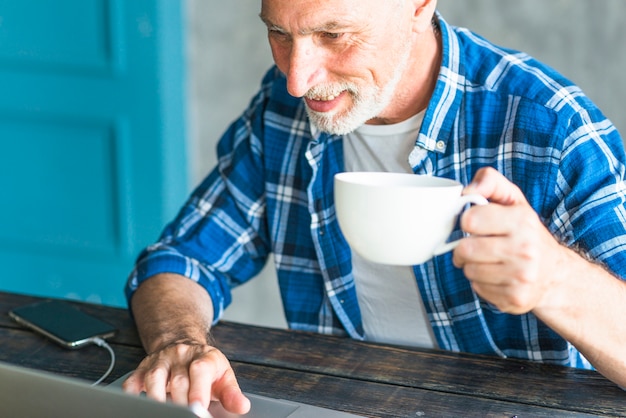 Equipaggi la tazza di caffè della tenuta facendo uso del computer portatile sullo scrittorio