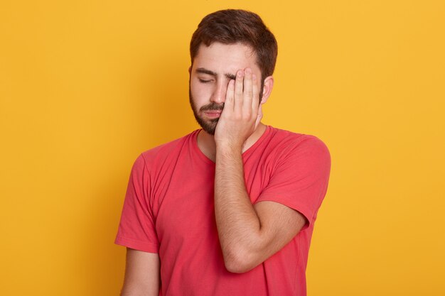 Esausto giovane con la barba lunga e la metà del viso con la mano, mentre in piedi contro lo studio giallo, sembra stanco, sentendosi depresso