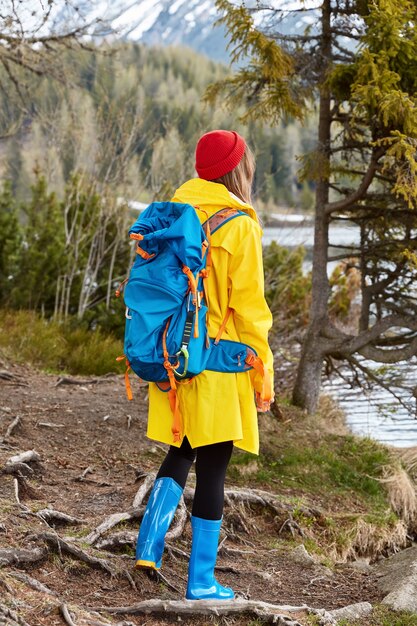 Escursionismo donna con zaino si trova in riva al lago, gode di vista nella natura, indossa un impermeabile giallo e stivali di gomma