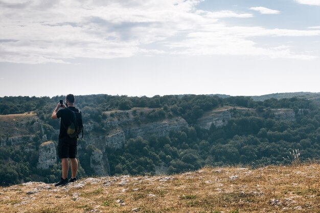 Escursionista maschio che scatta una foto di colline ricoperte di verde con il suo telefono sotto un cielo nuvoloso