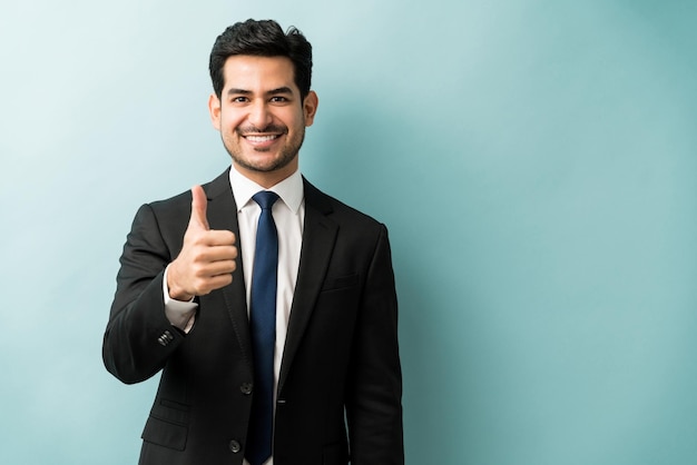 Esecutivo maschio ispanico sorridente che gesturing i pollici in su contro il fondo isolato