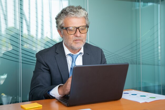 Esecutivo maturo serio che indossa vestito e occhiali, lavorando al computer in ufficio, utilizzando il computer portatile al tavolo