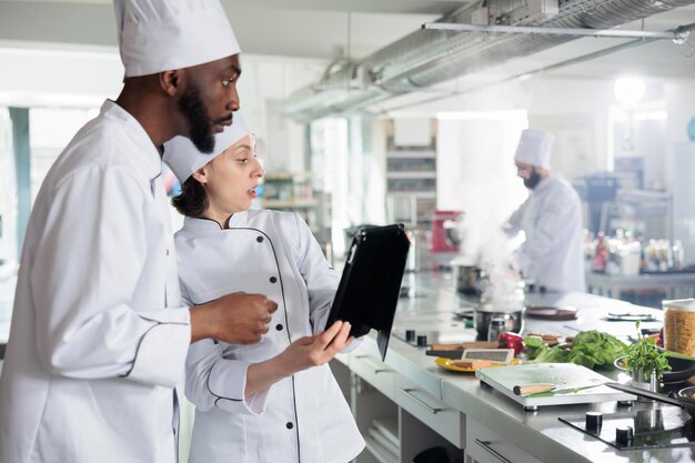 Esperti di cucina gourmet con tablet moderno alla ricerca di idee per piatti da tavola su Internet. Chef professionisti con dispositivo touchscreen portatile che preparano gli ingredienti per il servizio serale.