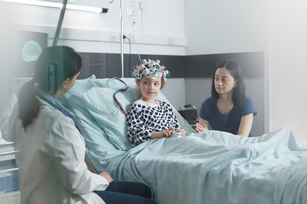 Esperto pediatrico della clinica che controlla i risultati della scansione EEG della ragazza malata mentre si trova nella sala di consultazione del reparto pediatrico. Medico esperto e madre ansiosa che discutono del trattamento e del periodo di recupero.