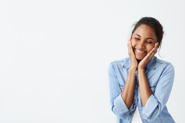 Espressioni ed emozioni del volto umano. Giovane donna afro-americana con i capelli neri e gli occhi chiusi, sorridendo con piacere, tenendosi per mano sulle guance, avendo un'espressione felice e calma del viso.