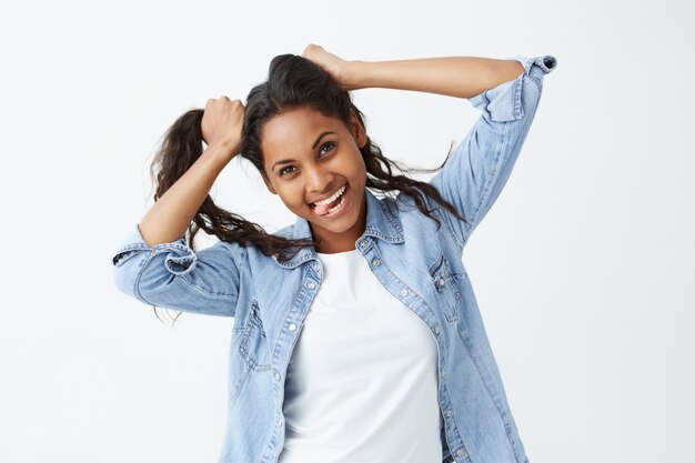 Espressioni ed emozioni del volto umano. Ritratto di giovane donna afro-americana giocosa in camicia di jeans, giocando con i suoi capelli, tenendo i capelli con entrambe le mani, con la bocca aperta in un sorriso gioioso.