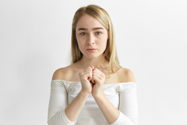 Espressioni facciali umane e linguaggio del corpo. Foto di elegante giovane femmina caucasica con sciolti capelli biondi in posa all'interno, tenendo due pugni serrati insieme, avendo a disagio nel profondo dei pensieri guardare