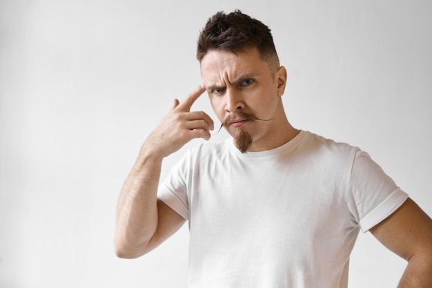 Espressioni facciali umane negative e linguaggio del corpo. Foto di moda giovane barbuto hipster con divertenti baffi a manubrio in posa in studio, essere arrabbiato, rotolare il dito indice alla tempia