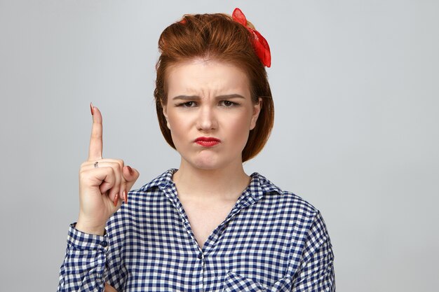 Espressioni facciali umane negative, emozioni e reazioni. Emotiva infelice giovane donna affascinante vestita in camicia a scacchi accigliato e imbronciato, avendo sconvolto sguardo scontroso, alzando il dito indice