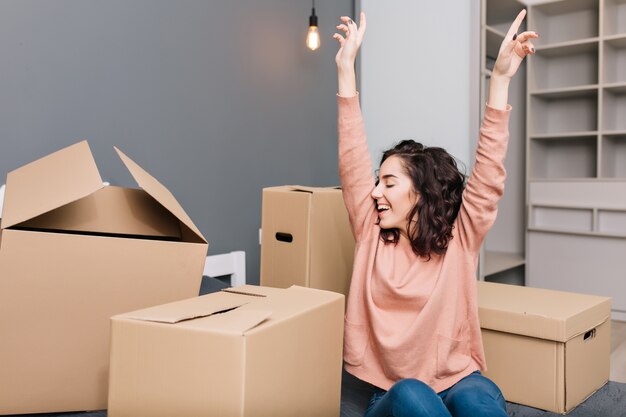 Esprimendo vivaci emozioni vere, positività della giovane donna graziosa con i capelli ricci castani corti sul letto circondano le scatole di cartone in appartamento moderno. Divertirsi in movimento, felicità nella nuova casa