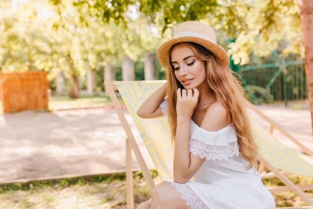 Estatica donna dai capelli lunghi con trucco elegante gode di una buona mattinata di sole seduti sulla sedia da giardino. Ritratto all'aperto di una ragazza spettacolare in barca a vela di paglia agghiacciante in chaise-long con gli occhi chiusi.