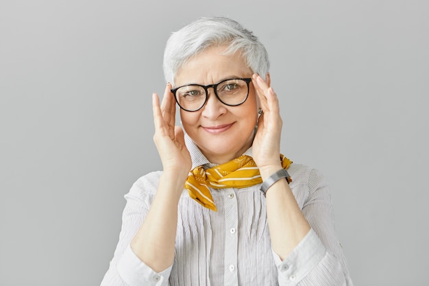 Età, ottica, occhiali e concetto di visione. Sorridente bello elegante pensionato femmina matura avente gioiosa espressione facciale, regolazione elegante occhiali in cornice nera, indossa una camicia e sciarpa