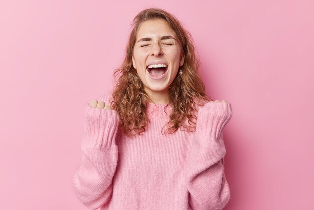 Euforica felicissima donna dai capelli lunghi tiene la bocca ampiamente aperta esclama di gioia stringe i pugni con trionfo celebra il successo indossa un maglione caldo isolato su sfondo rosa raggiunge l'obiettivo