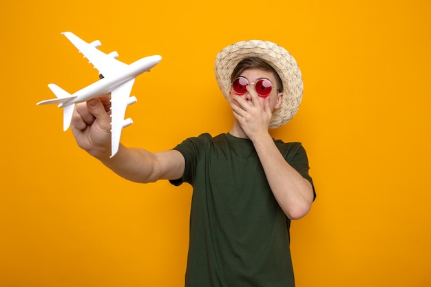 Faccia coperta spaventata con la mano giovane bel ragazzo che indossa un cappello con gli occhiali che tengono l'aeroplano giocattolo