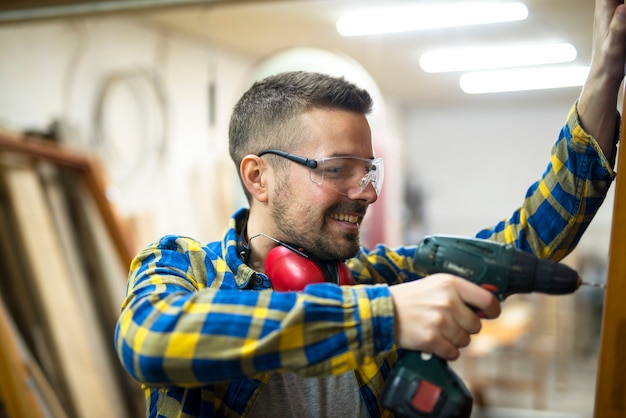 Falegname giovane lavoratore professionista con occhiali protettivi che tiene trapano e lavora al suo progetto in officina