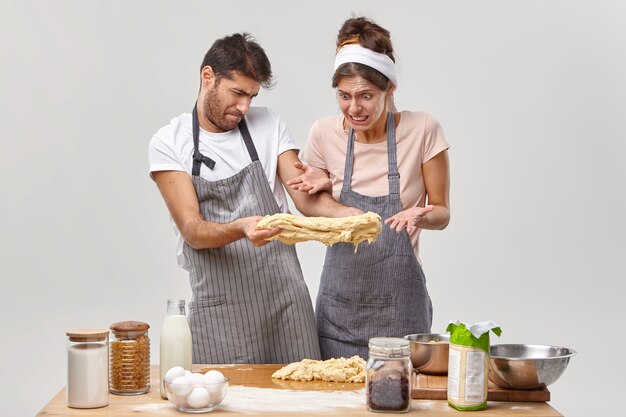 Fallimento e concetto di cucina. L'uomo e la donna delusi allungano la pasta cruda appiccicosa, hanno qualche problema con la pizza fatta in casa, un vero disastro in cucina, sguardo con facce disgustose, stoviglie sul tavolo