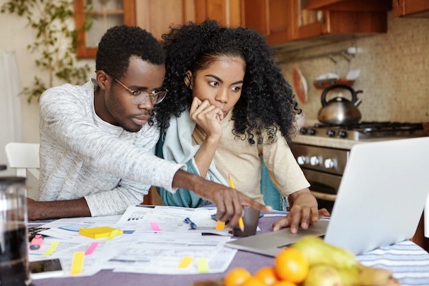 Famiglia africana di due persone seduto al tavolo nella loro cucina e pagare le bollette online utilizzando il computer portatile: uomo con gli occhiali che punta il dito indice contro lo schermo del notebook, spiegando qualcosa a sua moglie