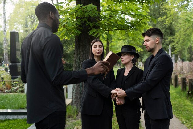 Famiglia al cimitero che si tiene per mano mentre il sacerdote legge la Bibbia