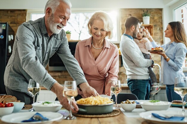 Famiglia allargata felice che pranza nella sala da pranzo. Il focus è sulla coppia matura che porta il cibo al tavolo.