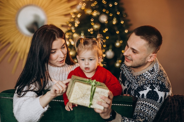 Famiglia con la piccola figlia che si siede dall'albero di Natale e che disimballa il contenitore di regalo