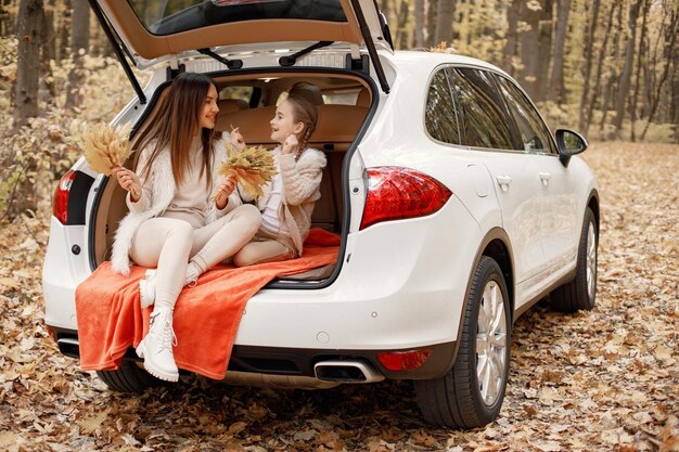 Famiglia felice che riposa dopo una giornata trascorsa all'aperto nel parco autunnale. La madre e la sua bambina seduta all'interno del bagagliaio di un'auto bianca. Madre e figlia che indossano abiti bianchi.