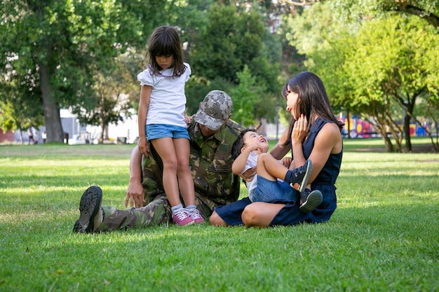 Famiglia felice che si siede sull'erba nel parco cittadino. Padre di mezza età caucasico in uniforme militare, madre sorridente e bambini che si rilassano insieme sul prato. Ricongiungimento familiare, fine settimana e concetto di ritorno a casa