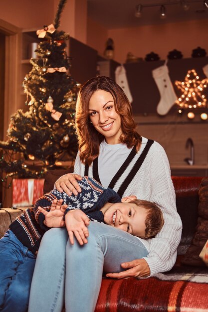 Famiglia felice. Mamma che abbraccia il suo bambino carino mentre è seduto sul divano nella stanza decorata durante il periodo natalizio.