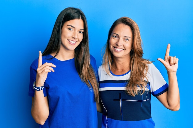 Famiglia ispanica di madre e figlia che indossano abiti casual su sfondo blu che punta il dito verso l'alto con un'idea di successo. uscito e felice. numero uno.