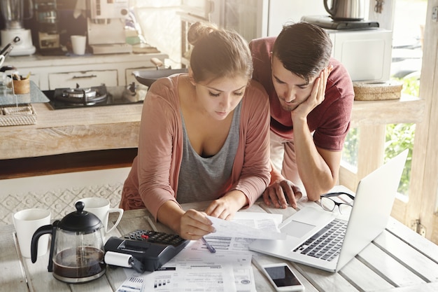 Famiglia stressata giovani che paga le bollette online usando il computer portatile. Documento preoccupato della tenuta della donna, calcolante le spese domestiche insieme a suo marito