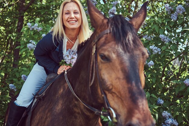 Fantino biondo affascinante felice che monta un cavallo marrone nel giardino fiorito.