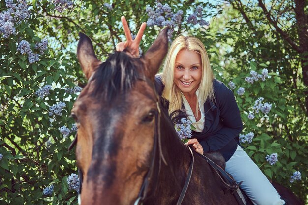 Fantino biondo affascinante felice che monta un cavallo marrone nel giardino fiorito.