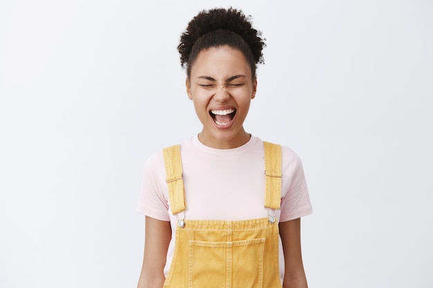 Far uscire le emozioni fa sentire bene. Incurante bella afroamericana con pelle liscia e pulita in tuta gialla, gridando con gli occhi chiusi, rilasciando lo stress, cercando di calmarsi sul muro grigio