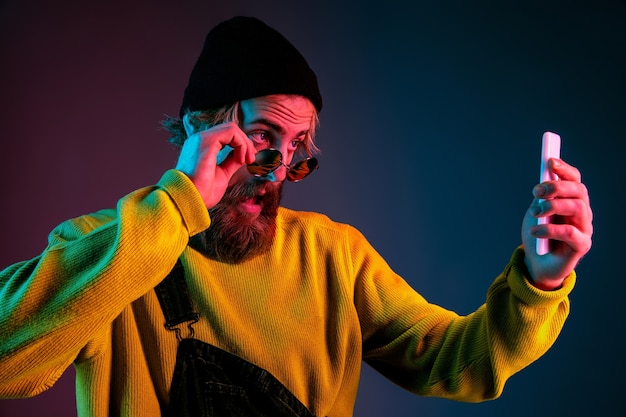 Fare selfie con gli occhiali. Ritratto dell'uomo caucasico sul fondo dello studio sfumato in luce al neon. Bellissimo modello maschile con stile hipster. Concetto di emozioni umane, espressione facciale, vendite, annuncio.