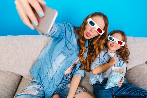 Fare selfie ritratto di momenti felici della vita familiare. Bella madre con lunghi capelli castani e piccola figlia che si diverte in occhiali 3D sul divano isolato su sfondo blu