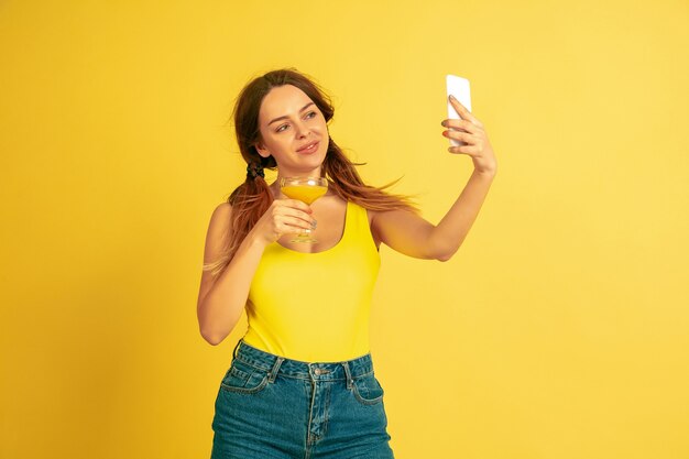 Fare selfie, vlog, sorridere. Ritratto della donna caucasica su sfondo giallo studio. Bellissimo modello femminile. Concetto di emozioni umane, espressione facciale, vendite, annuncio. Estate, viaggi, resort.
