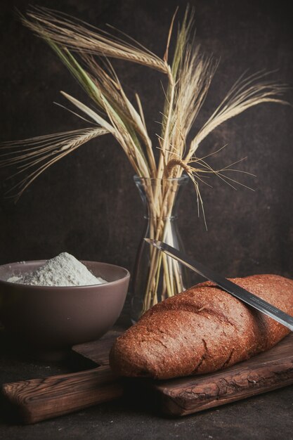 Farina di vista laterale in ciotola con grano, coltello e pane su marrone scuro. verticale