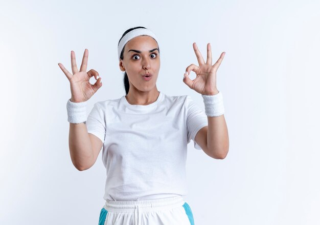 Fascia da portare e braccialetti da portare della donna sportiva caucasica sorpresa dei giovani che gesturing segno giusto della mano isolato su spazio bianco con lo spazio della copia