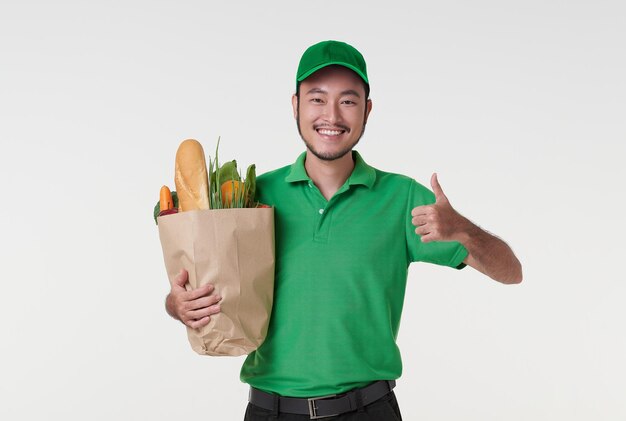 Fattorino asiatico che indossa in uniforme verde che tiene generi alimentari di sacchetti di carta per alimenti freschi e pollice in alto