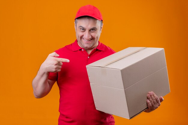 Fattorino che indossa l'uniforme rossa e cappuccio che tengono grande scatola di cartone che indica con il dito indice sorridendo sicuro sopra la parete arancio