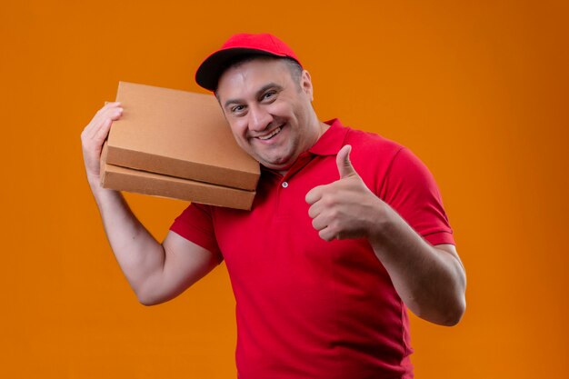 Fattorino che indossa l'uniforme rossa e cappuccio che tengono i contenitori di pizza positivi e felici che mostrano i pollici su sopra la parete arancio