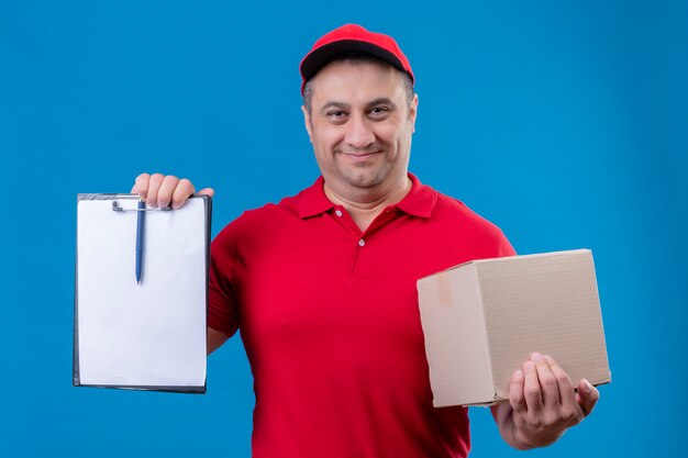 Fattorino che indossa l'uniforme rossa e cappuccio che tiene scatola di cartone che mostra lavagna per appunti con gli spazii in bianco che sembrano sorridere sicuro sopra la parete blu