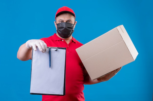 Fattorino che indossa l'uniforme rossa e cappuccio in maschera protettiva facciale che tiene appunti e scatola di cartone con la faccia seria sulla parete blu